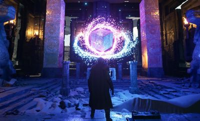 Záběr ze hry s postavou uprostřed, která se dívá na vznášející se magickou kostku v jasných živých barvách.