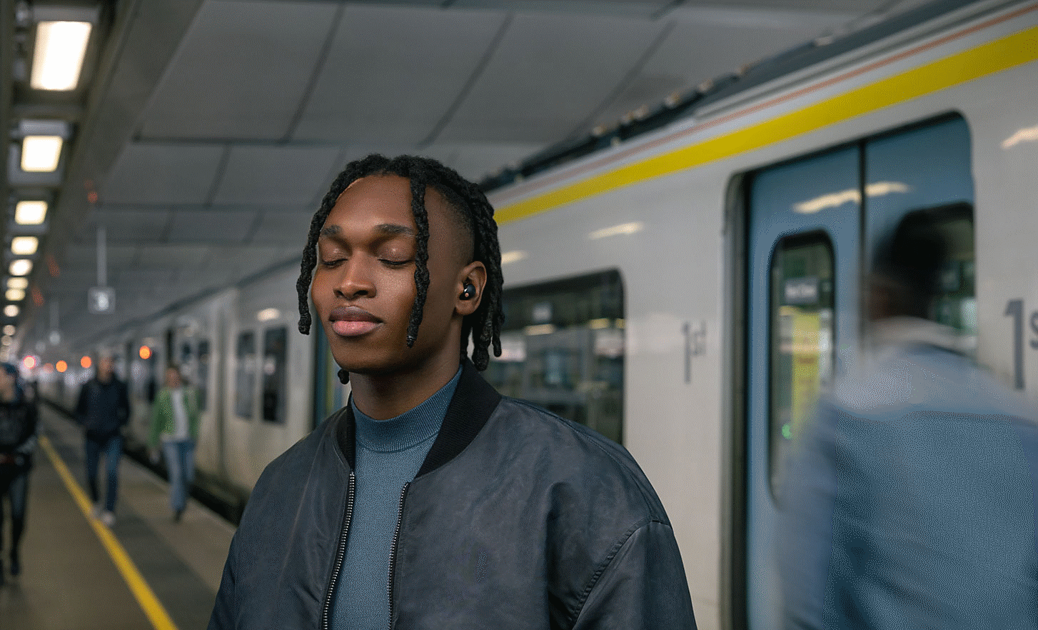Зображення людини із закритими очима, що стоїть на платформі поїзда в навушниках WF-1000XM5