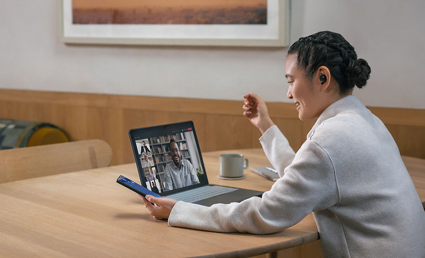 Imagen de una persona que usa los audífonos WF-1000XM5 sentada junto a una mesa, y mira su teléfono y laptop