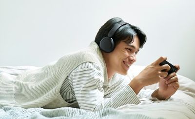 Mladý muž s černými sluchátky Sony WH-CH530 hraje na smartphonu v posteli