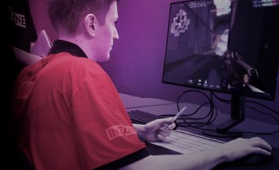 Ένα άτομο που φοράει κόκκινη μπλούζα με INZONE στο μανίκι και κάθεται μπροστά από μια οθόνη και ένα πληκτρολόγιο. Η οθόνη δείχνει μια σκηνή παιχνιδιού.