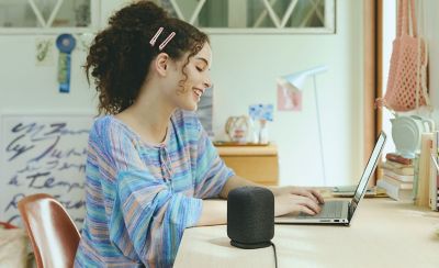 A young woman working at a laptop on a desk with the LinkBuds Speaker in the foreground.