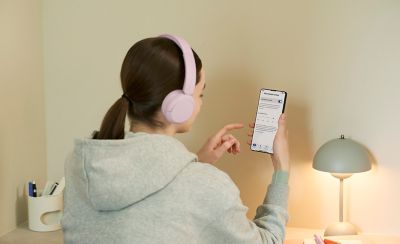Mladá žena používá smartphone k nastavení omezení hlasitosti na růžových sluchátkách Sony WH-CH530