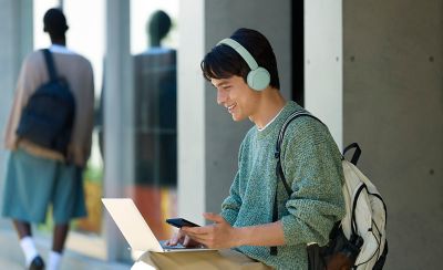Mladý muž venku s mátovými sluchátky Sony WH-CH530 pracující na notebooku a mobilním telefonu