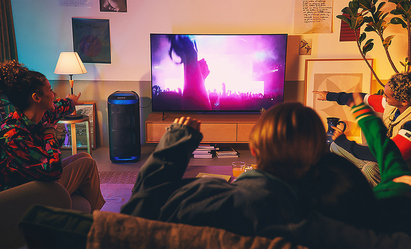 Kuva ihmisistä olohuoneessa katsomassa konserttia TV:stä, jonka vieressä on sinistä valoa hohtava SRS-XV800-kaiutin