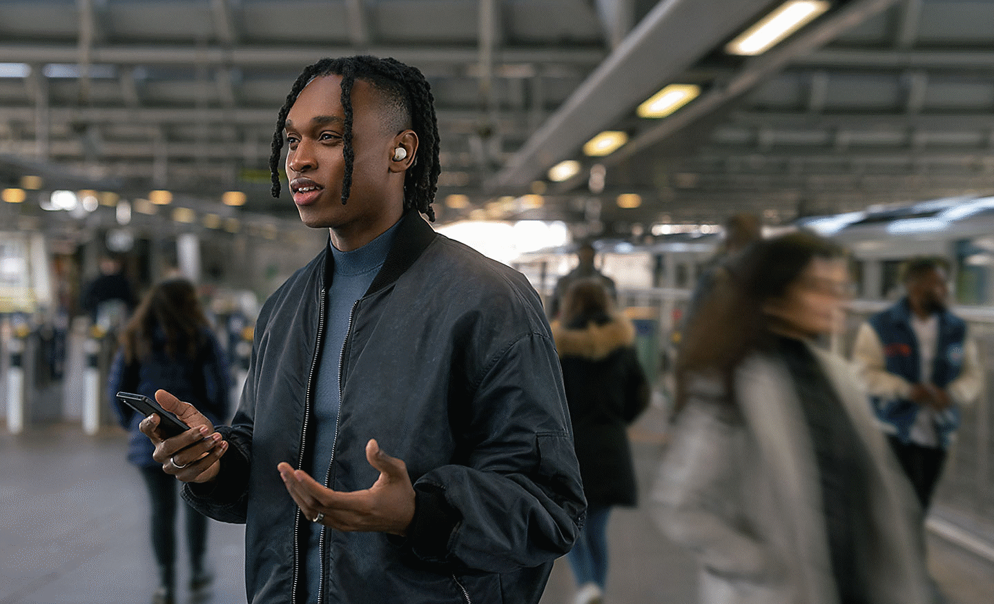 Imagen de una persona en una llamada, de pie en una estación de transporte público llena de personas mientras usa los auriculares WF-100XM5