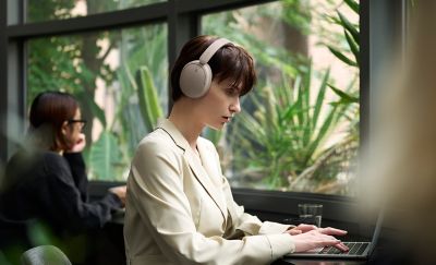 O femeie purtând o pereche de căști Sony WH-CH730N, modelul bej, lucrând la laptop într-o cafenea