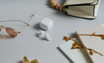 A pair of white LinkBuds Open earbuds and their charging case on a grey background with a pair of sunglasses, a notebook and a handbag arranged around them.