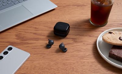 A pair of black LinkBuds Open earbuds and their charging case on a wood table with a smartphone, a laptop, a drink and a plate of cookies arranged around them.