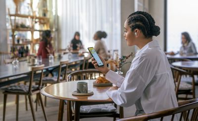 Image of a person sitting at a café wearing the WF-100XM5 headphones whilst looking at their phone