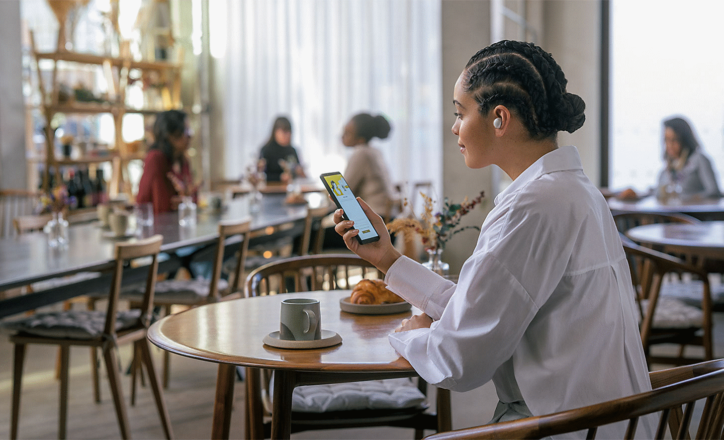 Imagine cu o persoană la cafenea, purtând căști WF-100XM5 în timp ce se uită la telefon