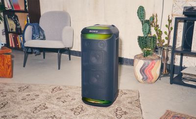 Image of the SRS-XV800 speaker with green ambient lightning sitting on a rug in a room with a chair and a cactus