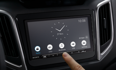 Afbeelding van de XAV-AX6050 in een dashboard met een klok en meerdere knoppen en iemand die een vinger voor het scherm houdt