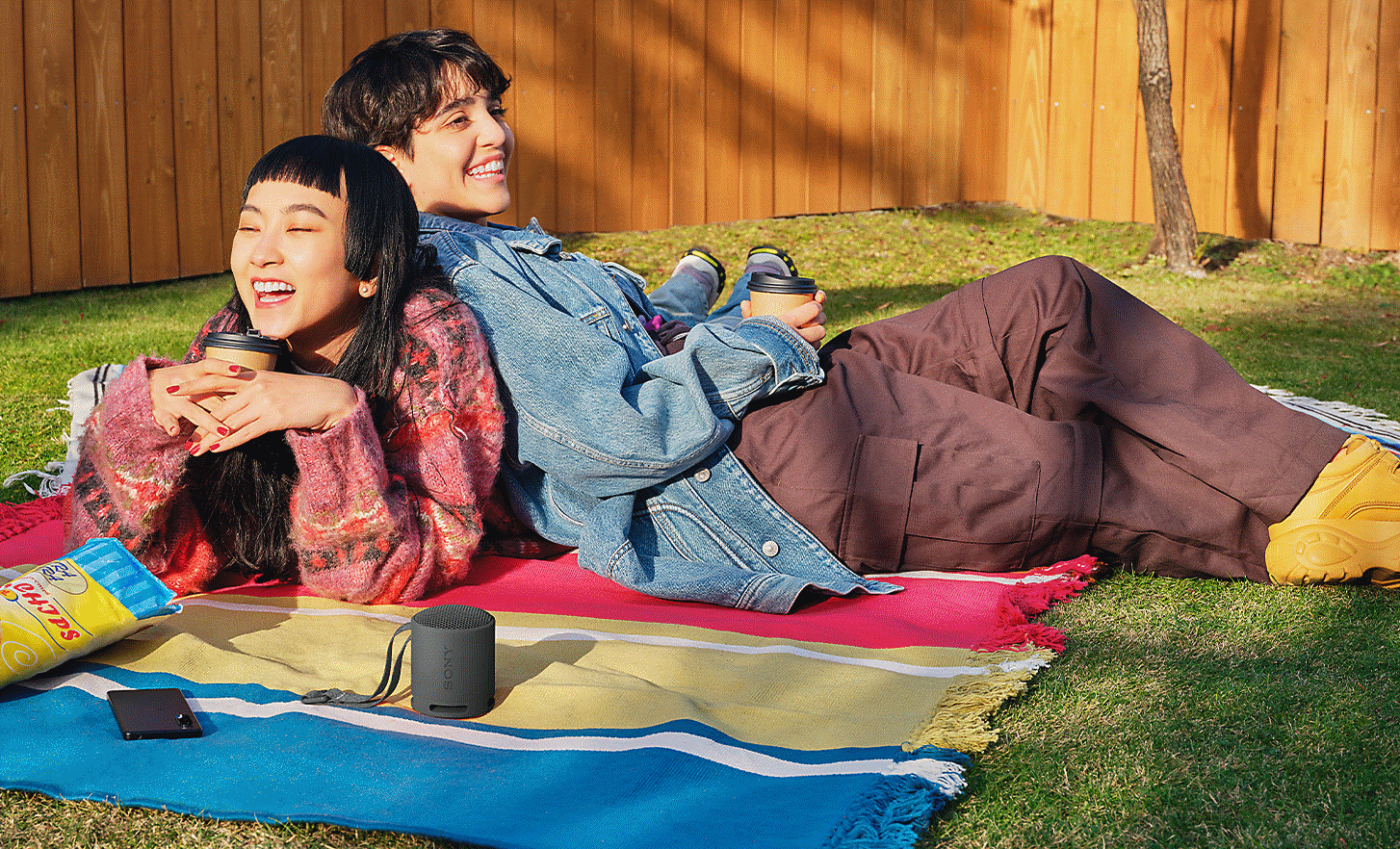 Obrázek dvou lidí ležících proti sobě na piknikové dece v zahradě, zatímco poslouchají černý reproduktor SRS-XB100