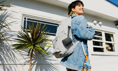 Image of a woman outside a white building wearing a backpack with a black SRS-XB100 speaker attached via its strap