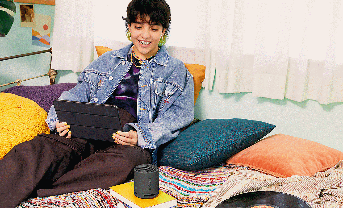 Abbildung einer Person mit einem Tablet auf dem Bett, die in einen auf einem Buch stehenden schwarzen SRS-XB100 Lautsprecher spricht