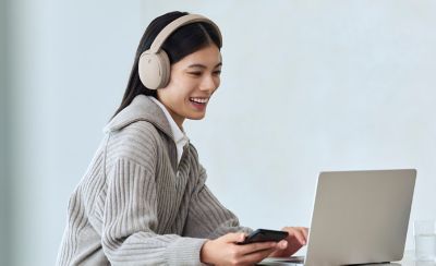 O femeie purtând o pereche de căști Sony WH-CH730N, modelul bej, folosind telefonul și laptopul