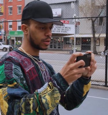 Мужчина в черной бейсболке держит черный смартфон на фоне сетчатого забора и улицы.