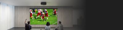 Deux hommes et une femme regardent une partie de football à l’aide d’un projecteur et d’un grand écran installé au mur.