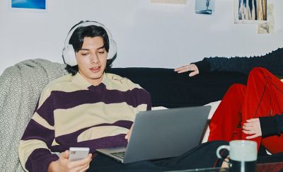 En ung person sitter vid sin bärbara dator med en telefon i handen och har på sig vita ULT WEAR-hörlurar.