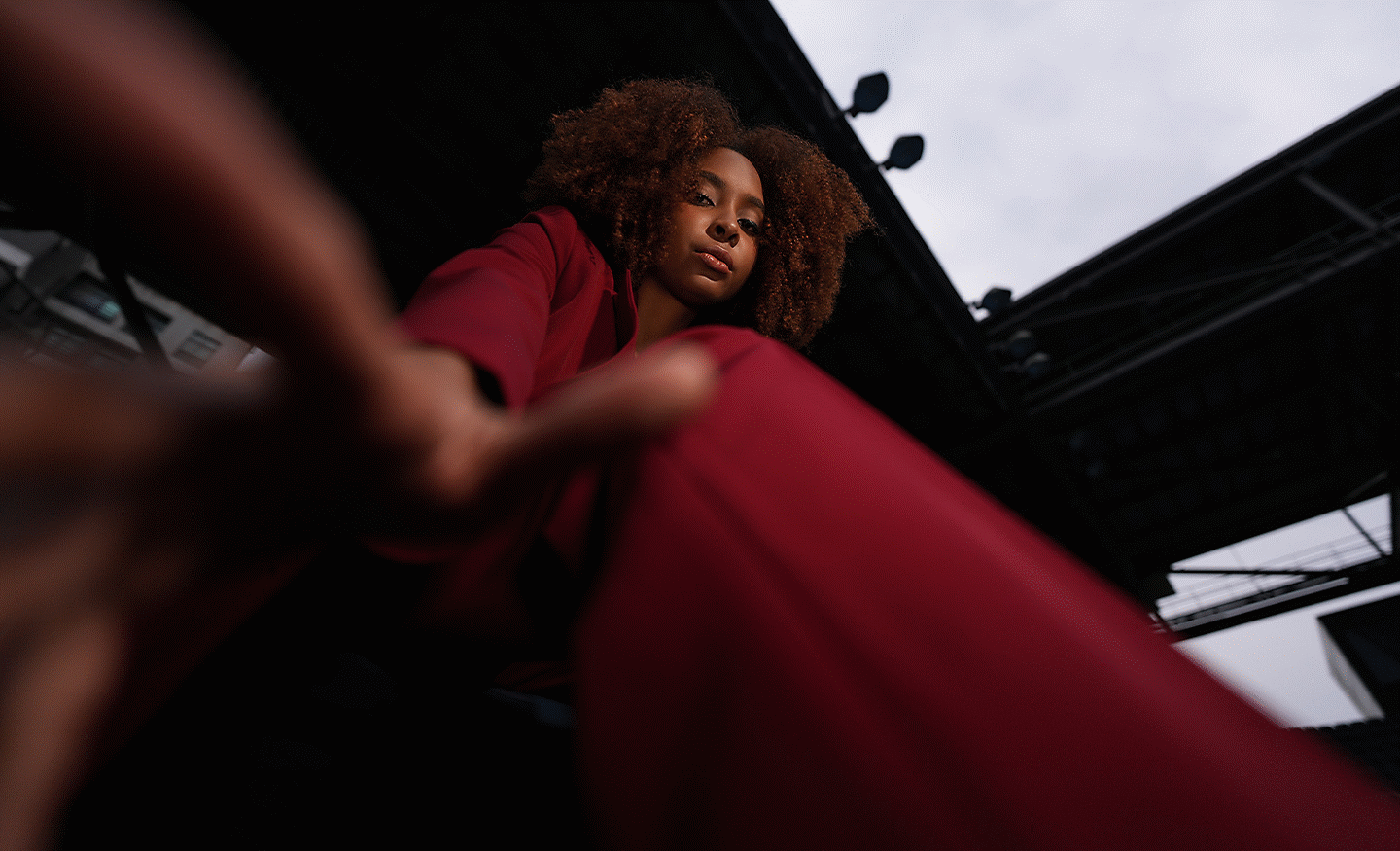 Low-angle portrait of a woman in a red pantsuit reaching toward the camera against an urban backdrop.