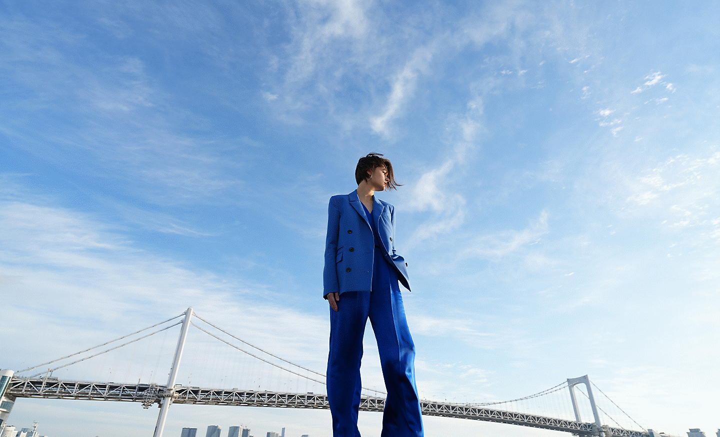 Full-body portrait of a person in a blue suit standing outdoors under a wide blue sky with a bridge in the background.