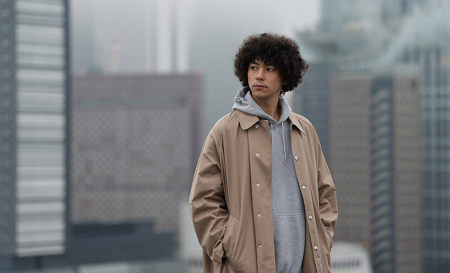 Portrait of a man wearing a beige coat standing on a rooftop with a city skyline in the background.