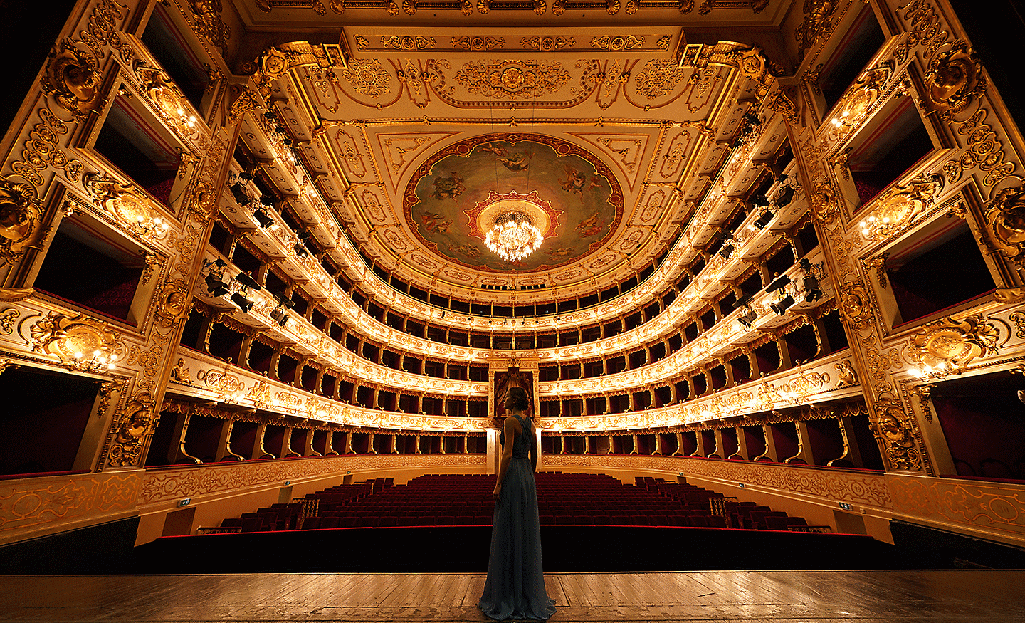 Symmetrical interior view of an ornate opera house with golden balconies and a person standing on the stage.