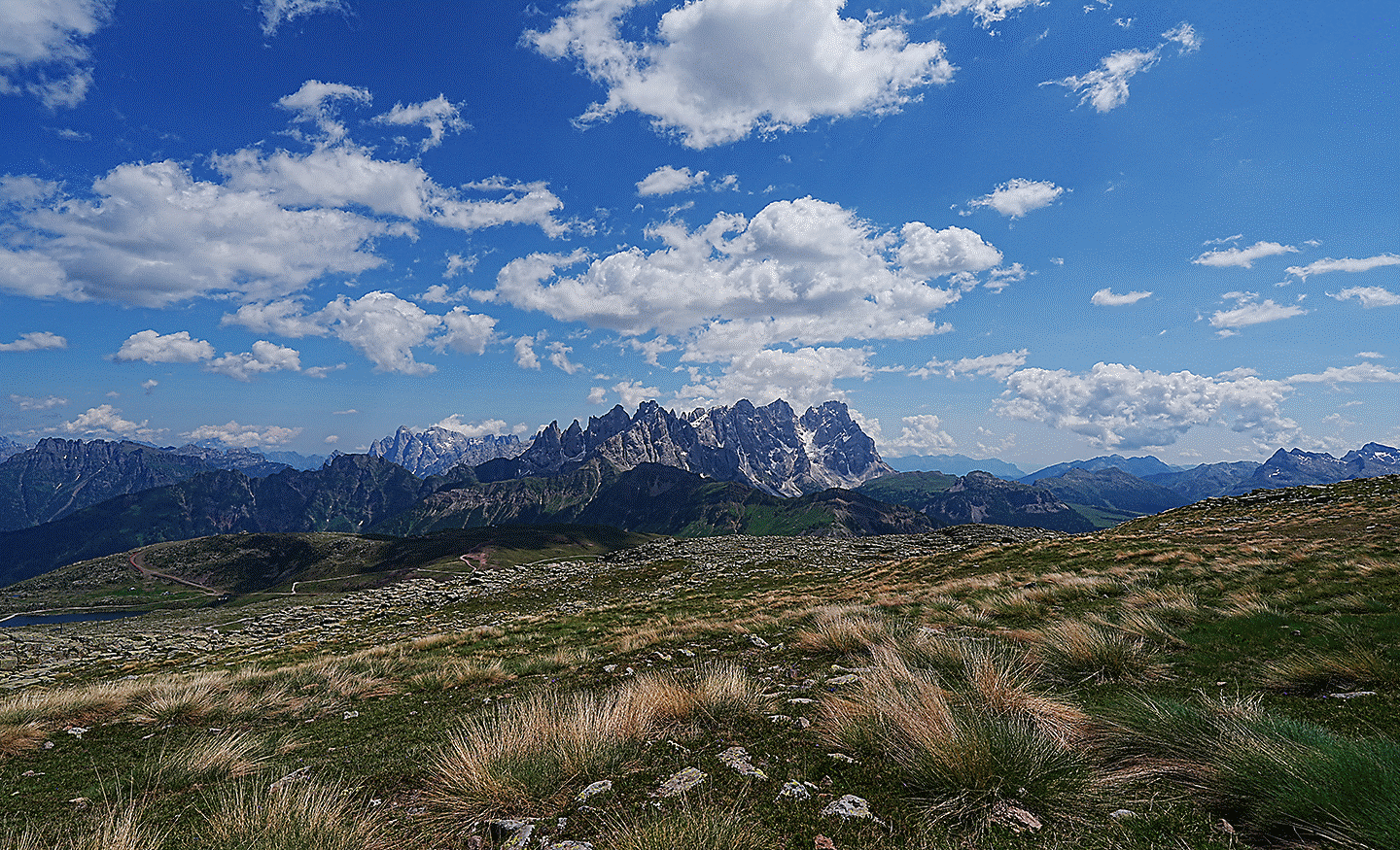 Wide landscape view of mountains under a blue sky with scattered clouds and grassy terrain in the foreground.