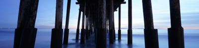 Symmetrical view from under a pier at dusk, with rows of wooden pillars leading toward the horizon over calm water.