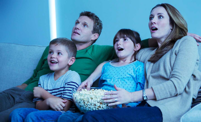 Une famille de quatre personnes est assise côte à côte sur un canapé tenant un bol de pop-corn, avec des expressions excitées sur leurs visages