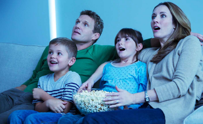 Eine vierköpfige Familie sitzt eng beieinander auf einem Sofa mit einer Schüssel Popcorn in der Hand und begeisterten Gesichtsausdrücken