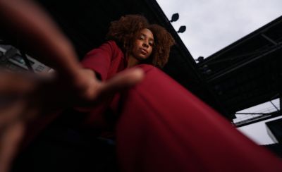 Low-angle portrait of a woman in a red pantsuit reaching toward the camera against an urban backdrop.