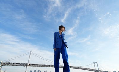 Full-body portrait of a person in a blue suit standing outdoors under a wide blue sky with a bridge in the background.