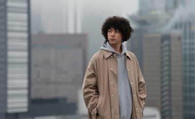 Portrait of a man wearing a beige coat standing on a rooftop with a city skyline in the background.