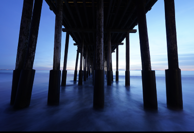 Symmetrical view from under a pier at dusk, with rows of wooden pillars leading toward the horizon over calm water.