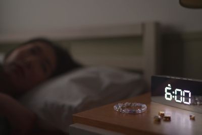 Hình ảnh có chiếc đồng hồ chỉ 6:00 trên kệ trong tiền cảnh và một người phụ nữ đang ngủ ở hậu cảnh