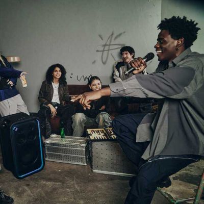 Un hombre participando en una batalla de rap con el ULT TOWER 7 y ULTMIC1 en un garaje, mientras sus amigos observan y disfrutan de la actuación.