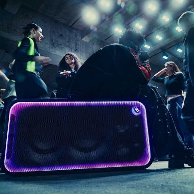 Un hombre sentado sobre el ULT TOWER MAX mientras la gente disfruta de la música a su alrededor en un ambiente de discoteca enérgico.