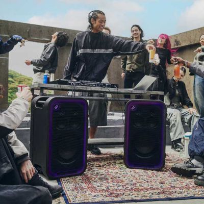 En una fiesta en una azotea, hay un DJ haciendo una presentación mientras la gente a su alrededor disfruta de la música, con dos parlantes ULT TOWER 7 en primer plano.